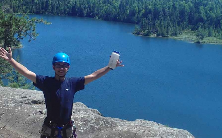 a person wearing rock climbing gear stands on a cliff above blue water and raises their hands in celebration